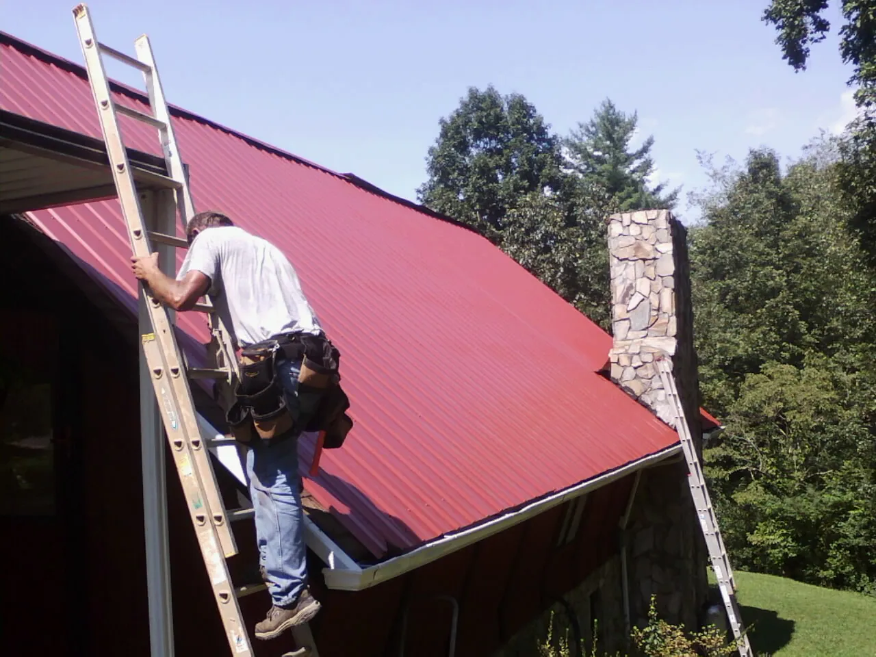Roofer on a ladder beside a red metal roof and stone chimney — Soffit & Fascia Repair by Roofing Brothers in Findlay